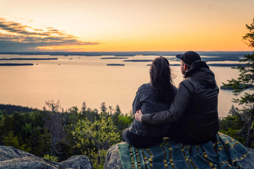 Maisemakuva Ukko-Kolilta auringonlaskussa. Parikunta istuu kalliolla ja katselee maisemaa.