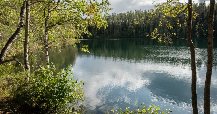 Kuvassa kesäinen järvi, johon heijastuu sinisiä pilviä. Ympärillä vihreää puustoa.