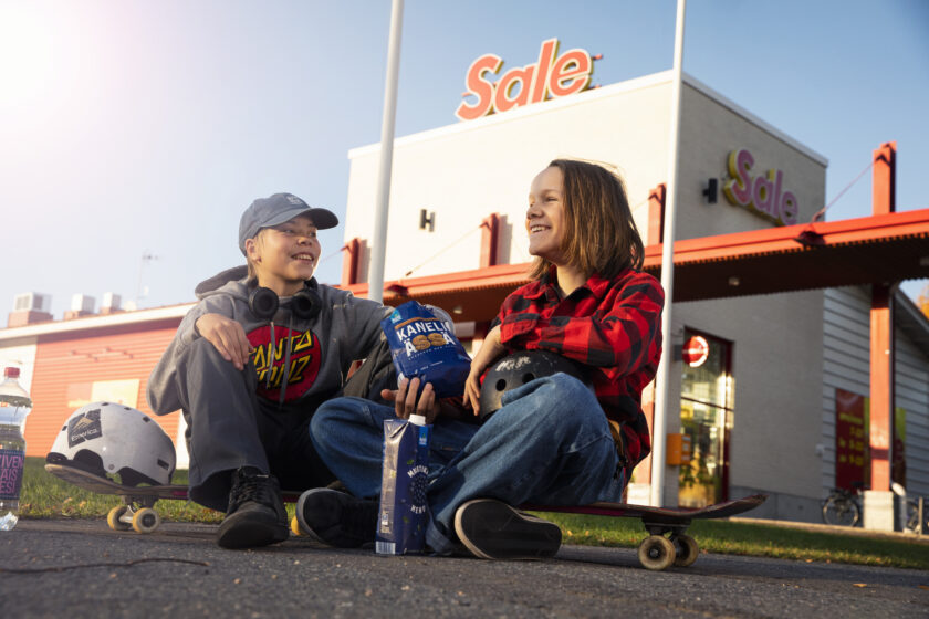 Taustalla valkoinen Sale-rakennus, etualalla skeittilautojen päällä istuu kaksi hymyilevää lasta.