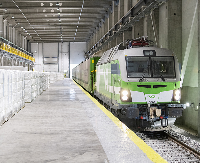 VR:n vihreä-valkoinen juna pysähtyneenä harmaan tehdasrakennuksen junanlastauslaiturilla.