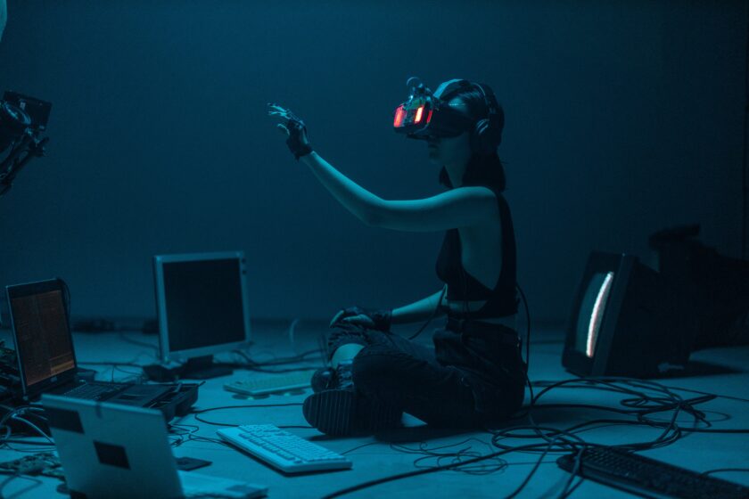 Tumman sininen, pimeä tunnelma. Huone jonka keskellä istuu nuori tyttö VR-lasit päässä. Käsi on nostettuna ylös. Ympärillä lattialla tietokonenäyttöjä ja johtoja.
